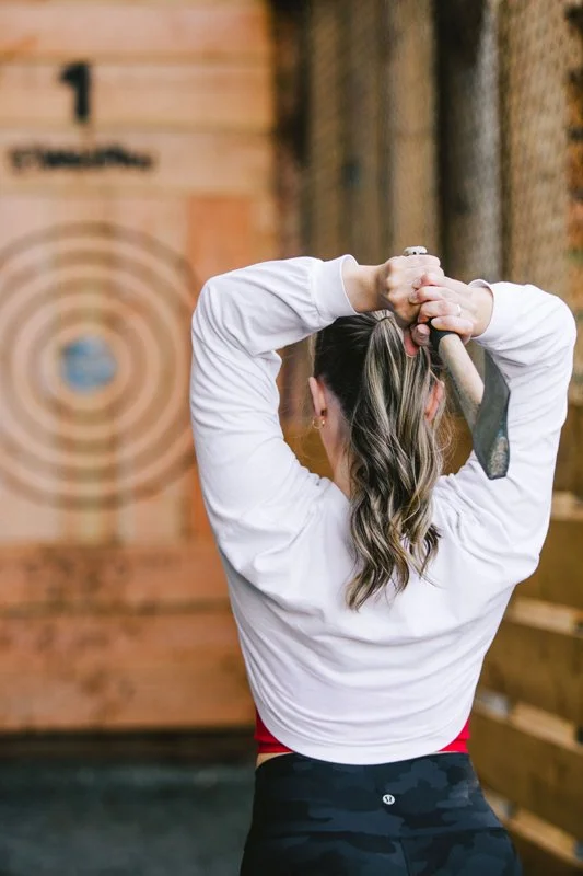 A woman with long, wavy hair tied in a ponytail, wearing a white long-sleeve shirt and black camouflage workout pants, is in an axe-throwing venue, preparing to throw an axe at a target.