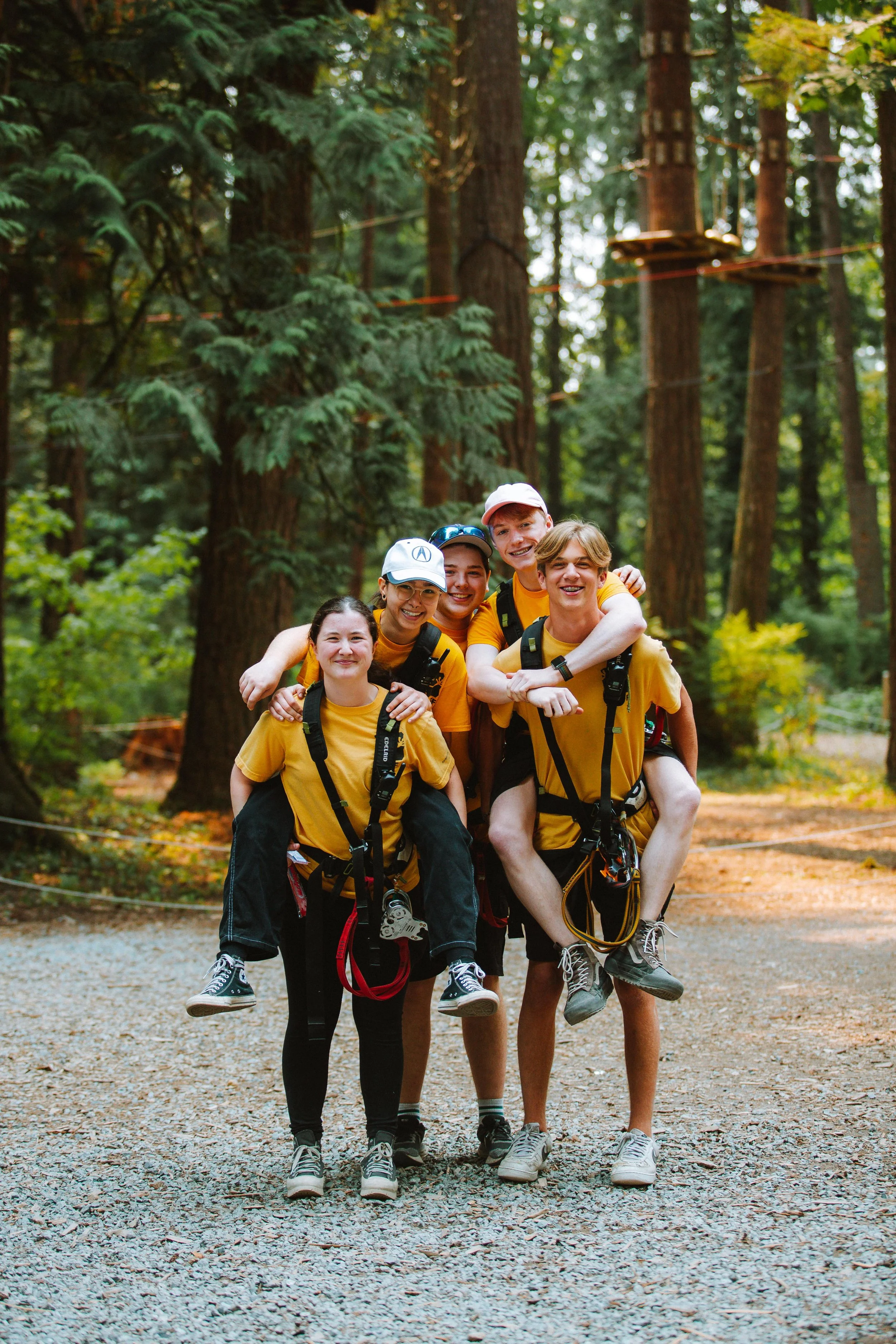 Group of five young friends in yellow shirts enjoying a group activity in a forest with trees, some of them are giving piggyback rides while smiling.