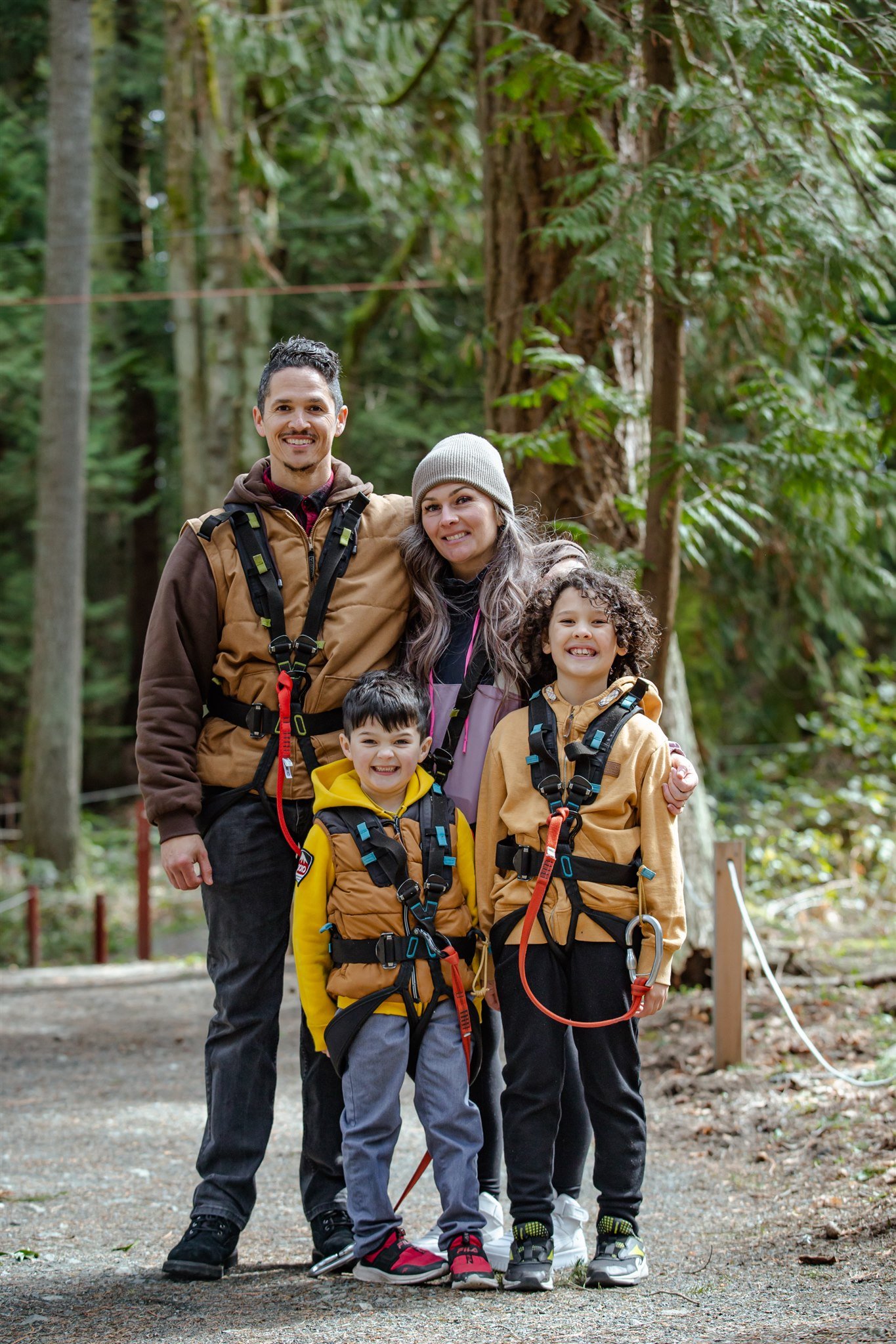 A family of four stands together in a forest, smiling, with hiking gear on, surrounded by tall trees.