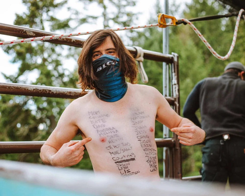 A shirtless person with shoulder-length hair, wearing a face mask, pointing to their chest with handwritten names written on their skin.