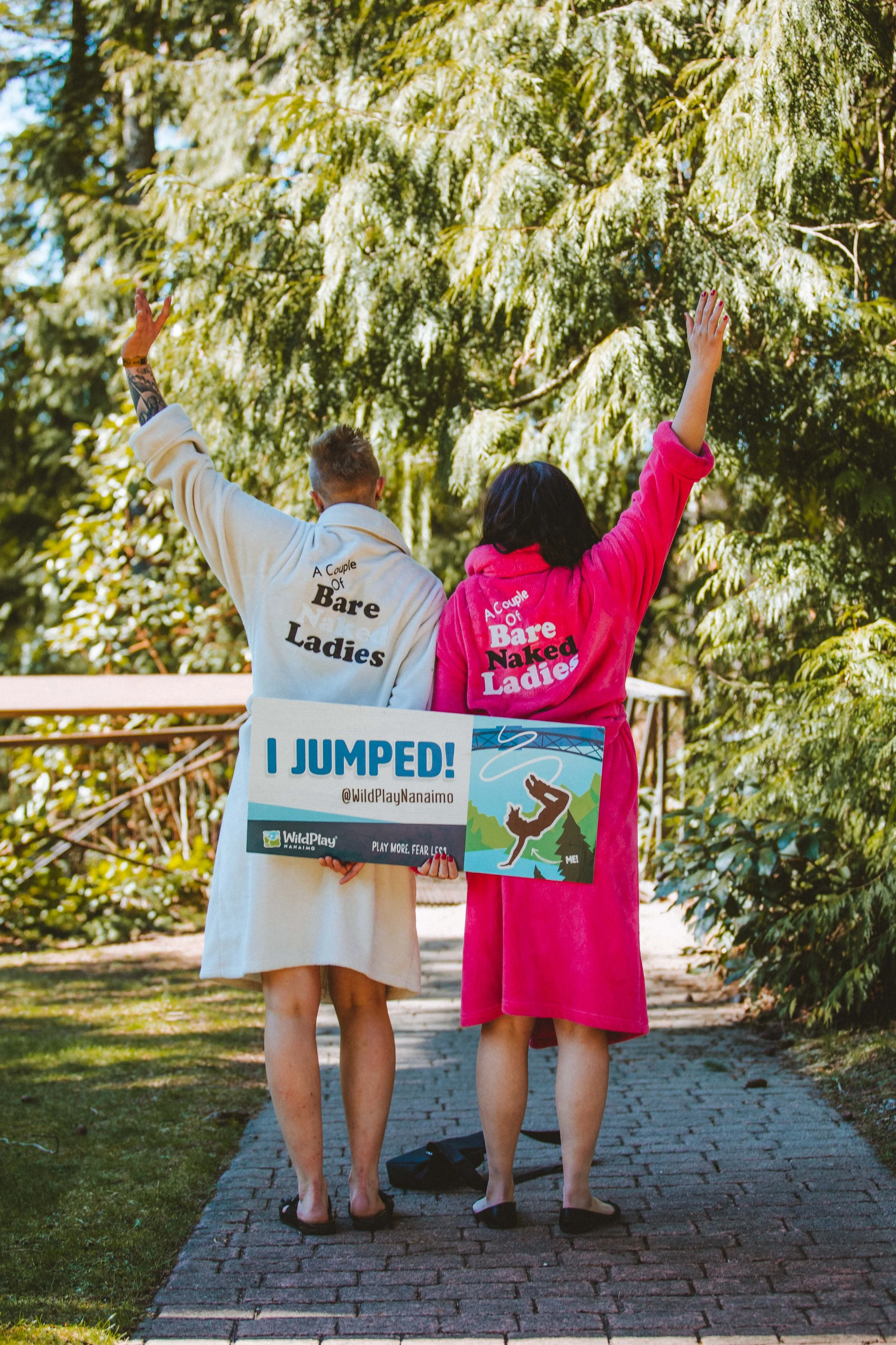 Two women standing on a brick pathway with their backs to the camera, wearing robes that say "A Couple of Bare Ladies" and holding a sign that says "I JUMPED! @WildPlayNanaimo." They are outdoors among lush green trees, celebrating their jump.