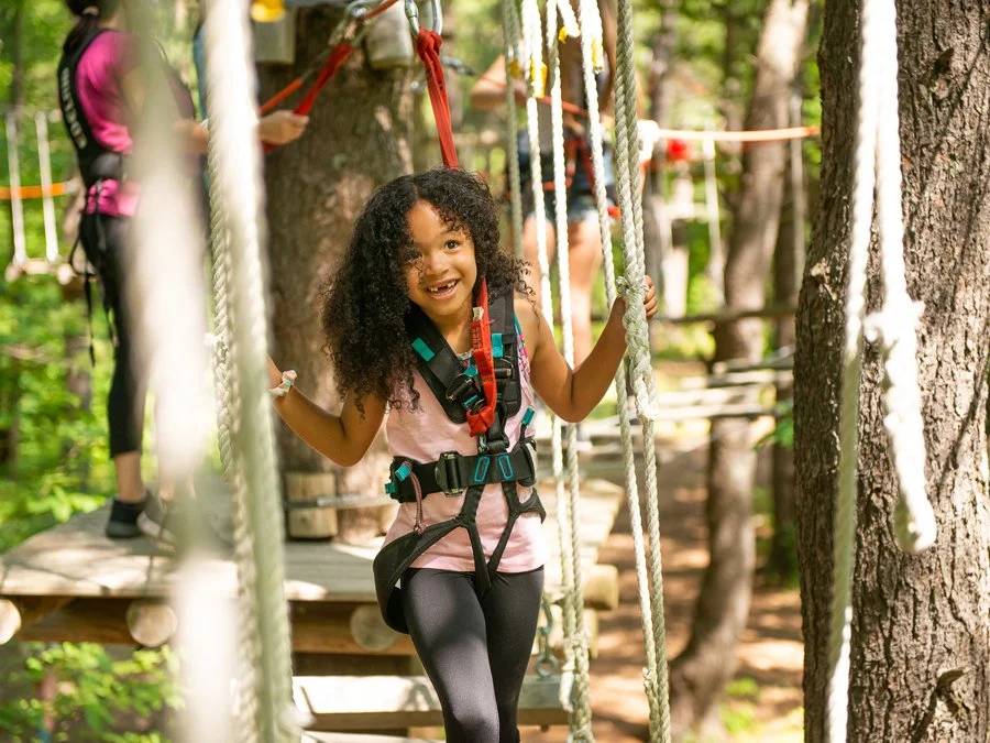 A young girl smiling and wearing a safety harness while walking on a rope bridge in a forested outdoor adventure park.