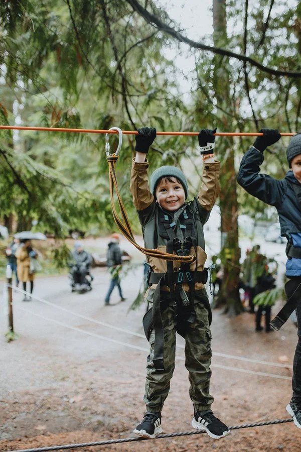 Young boy wearing a gray beanie and camouflage pants, smiling while hanging from a safety rope in a forested area during an outdoor adventure or zipline activity.