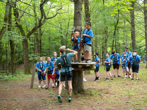 Group of children in a forest during a summer camp, some climbing a tree platform with a canopy of green leaves, supervised by a guide. 