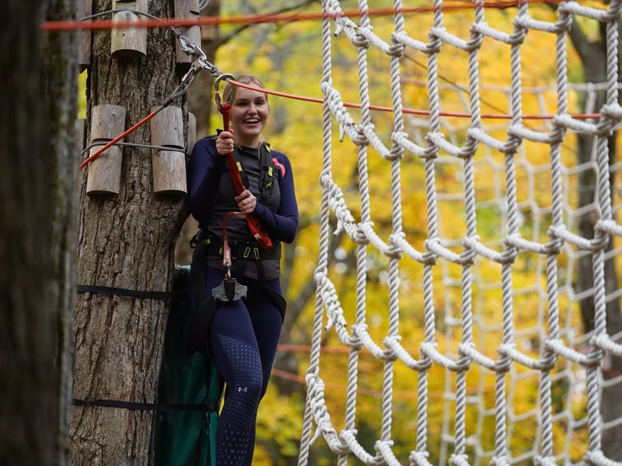 A woman smiling on a rope course in a forest with autumn leaves, wearing safety gear including a harness and helmet, holding a safety rope, and standing next to a large tree with a netted obstacle course.