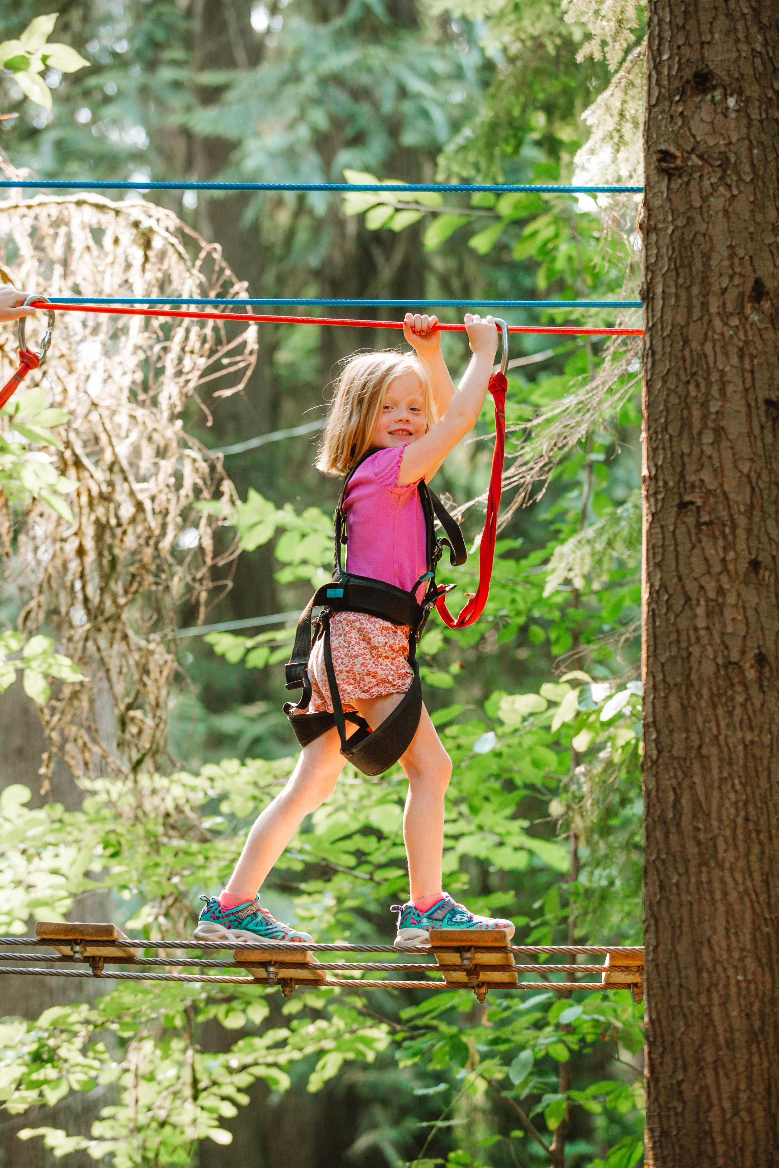 A young girl with blonde hair wearing a pink shirt, shorts, and hiking shoes, is walking on a wooden and rope bridge in a forest. She is holding onto a red safety handle and smiling.