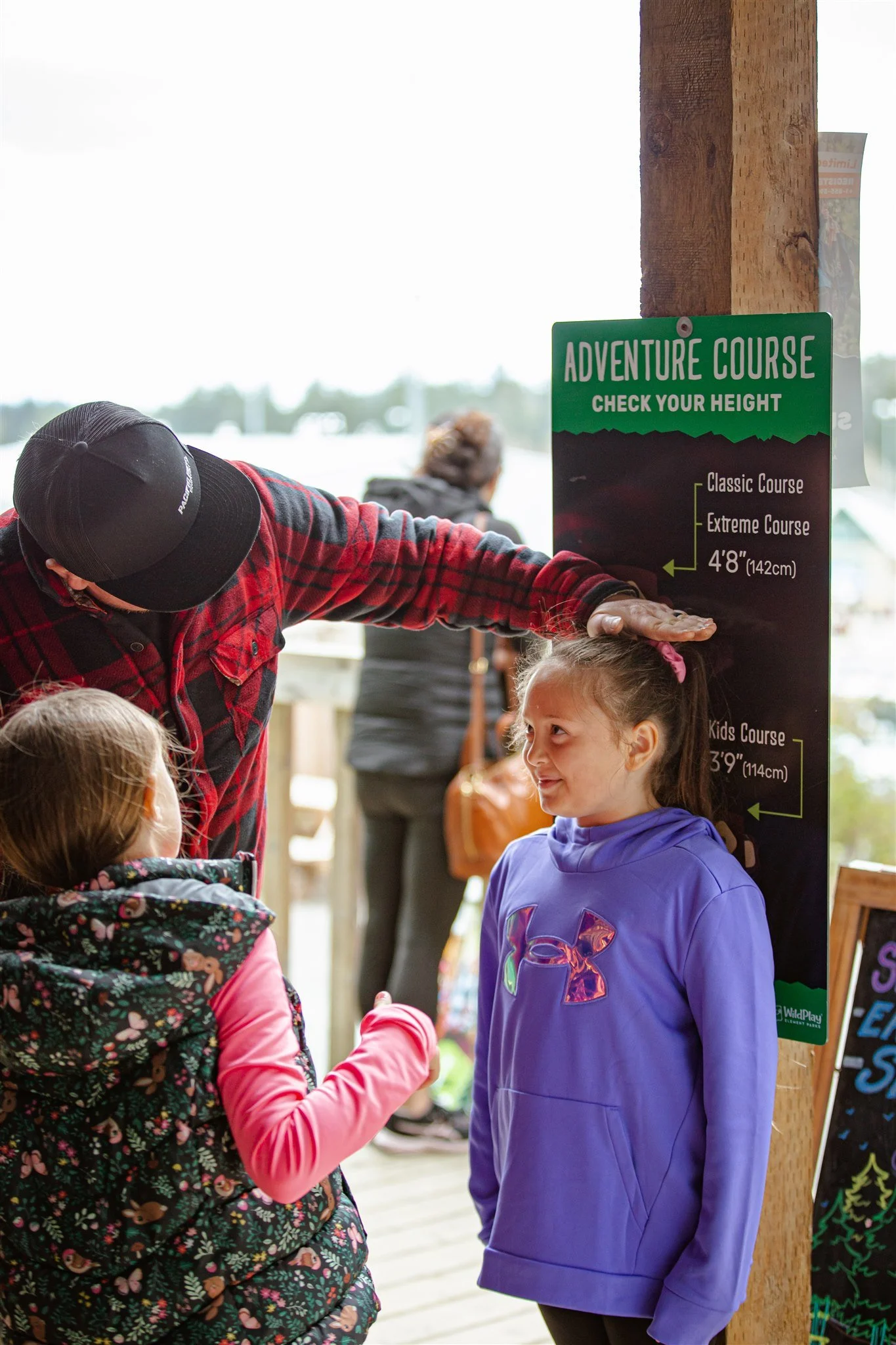 A man in a black cap and red plaid shirt measures the height of a young girl in a purple hoodie at an adventure course check-in sign.