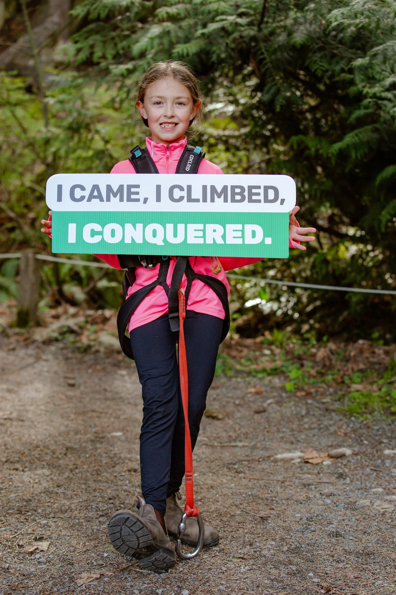 A young girl standing on a wooded trail holding a sign that reads, 'I CAME, I CLIMBED, I CONQUERED,' dressed in outdoor clothing and hiking boots.