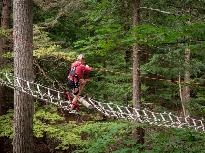A woman crossing a rope bridge in a forest, wearing a red jacket and harness.