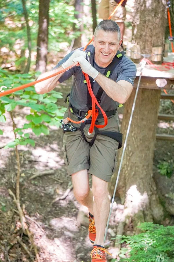 A man wearing shorts, a t-shirt, and hiking shoes is balancing on a slackline in a forest. He is smiling and holding a safety handle with his right hand.