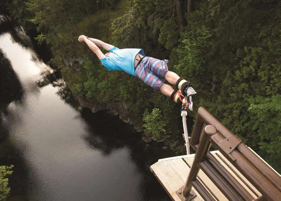 A person lying on a safety harness on a platform by a waterfall, with trees and water below.