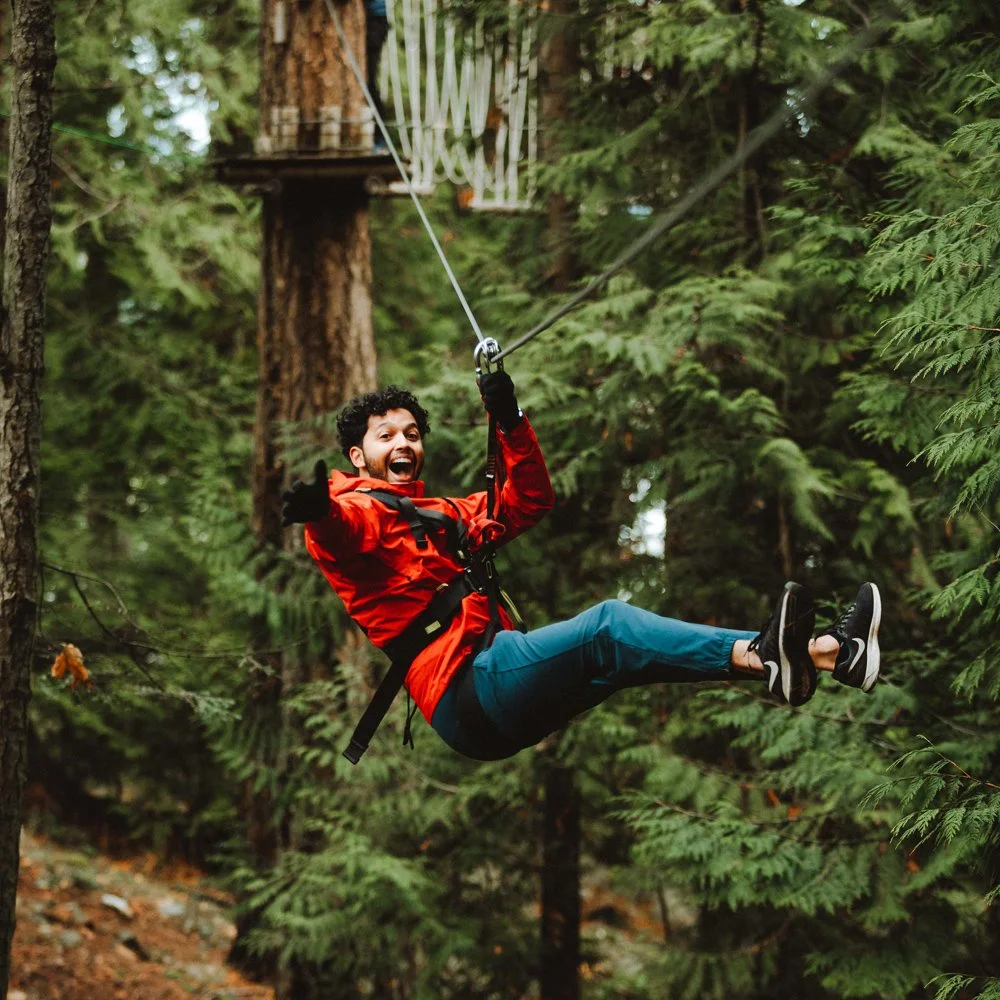 A man with a curly beard and hair, wearing a red jacket, black gloves, and black shoes, is smiling and enjoying a ziplining adventure through a forest with tall green trees.