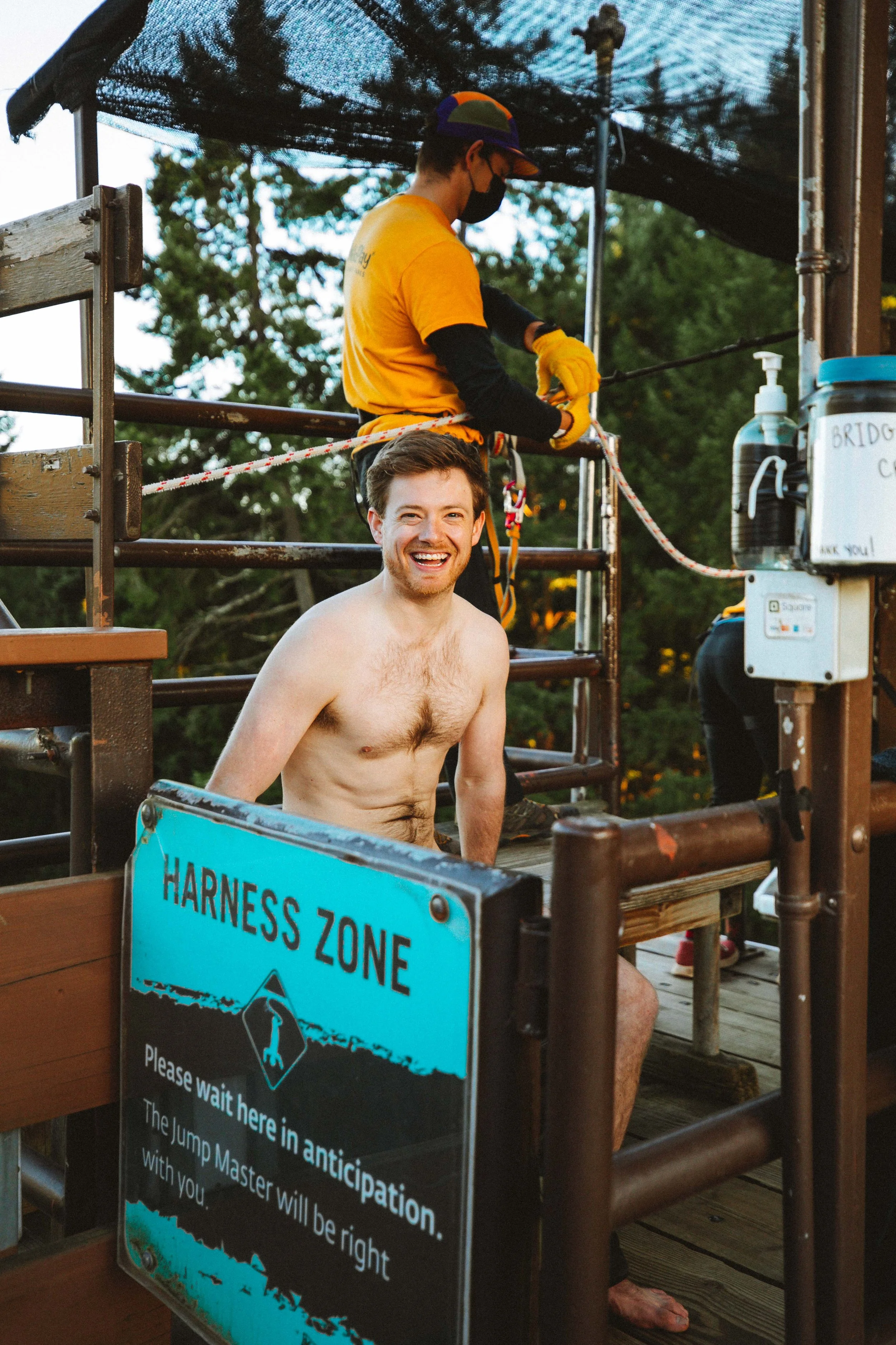 A shirtless man smiling and sitting at a water park or amusement park, with a sign reading 'Harness Zone' in front of him. In the background, a worker is preparing or securing a harness, wearing a face mask, helmet, and gloves.