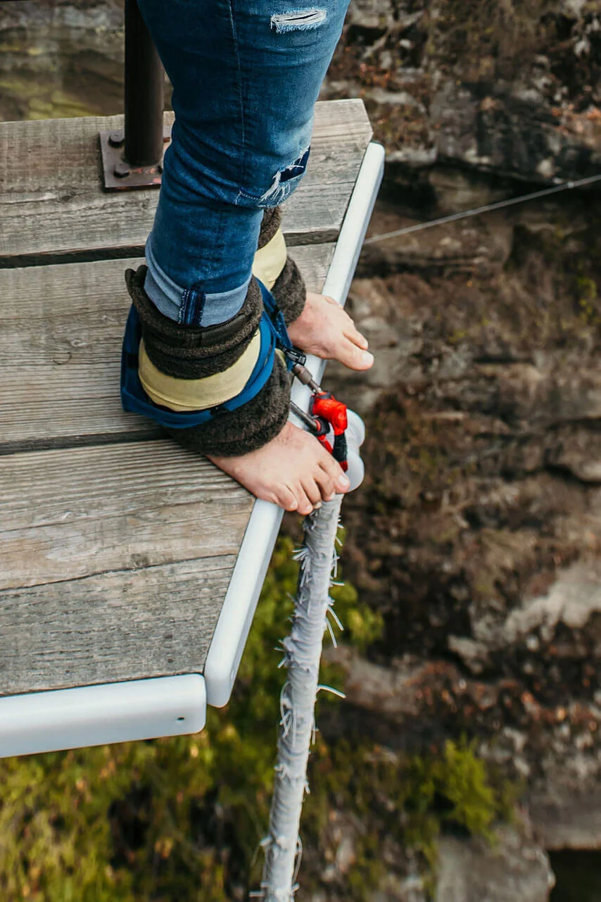 Person standing barefoot on a narrow wooden platform at the edge of a cliff, wearing ripped jeans, with a safety harness attached to the platform, holding onto a white rail, overlooking a rocky landscape.