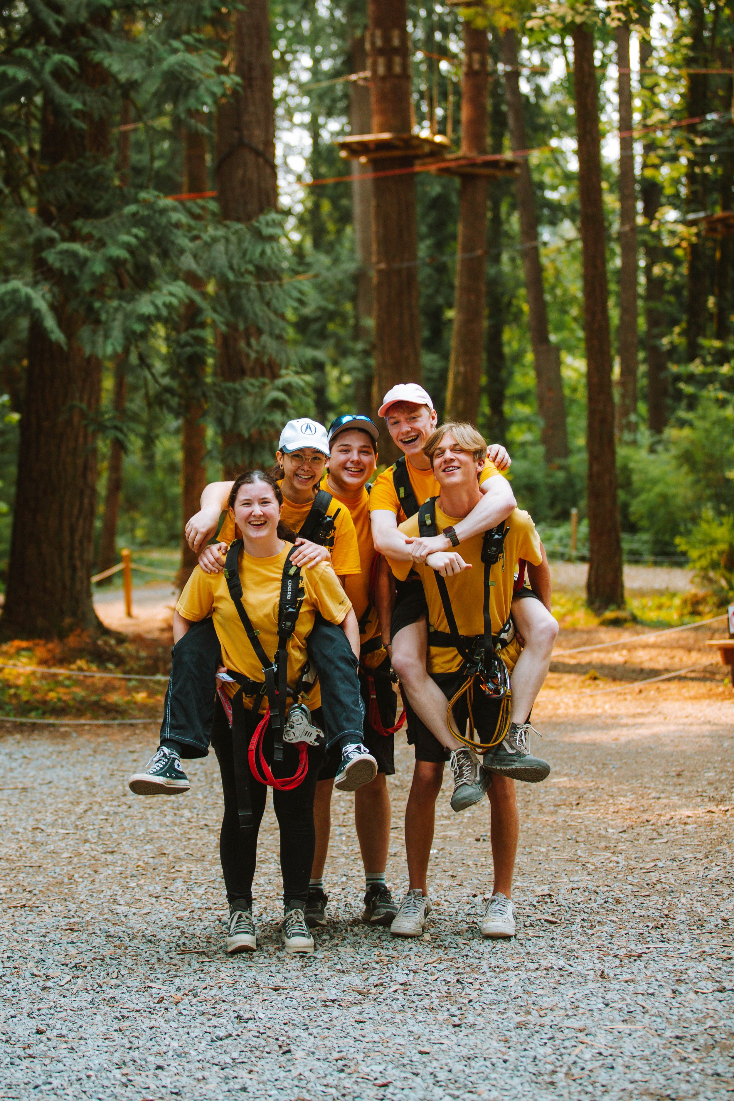 Group of five young people smiling and posing in a forest, with two of them piggybacking on others, wearing yellow shirts and harnesses, in a treetop adventure park.