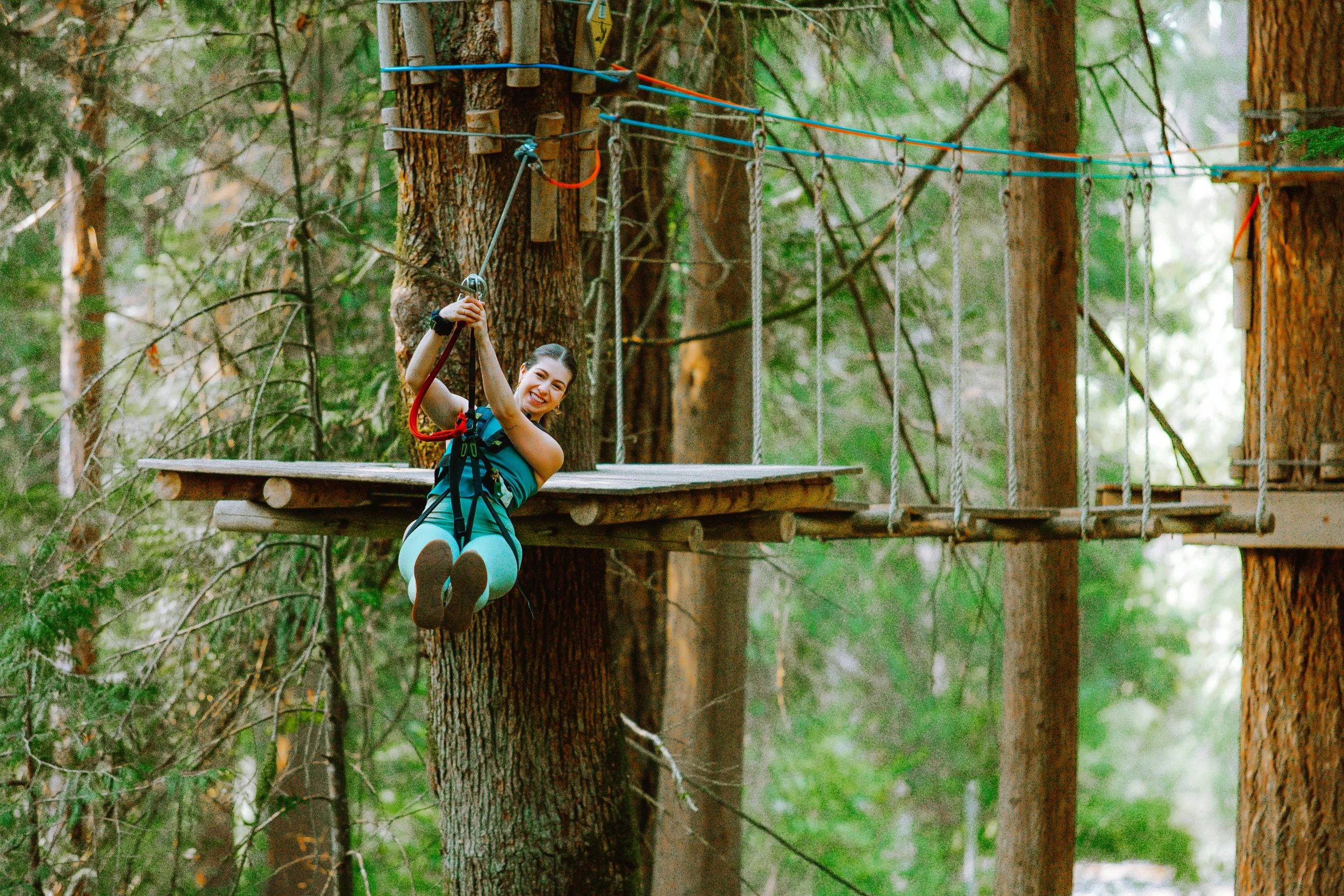 A young woman with a safety harness zip-lining through a forest with tall trees.