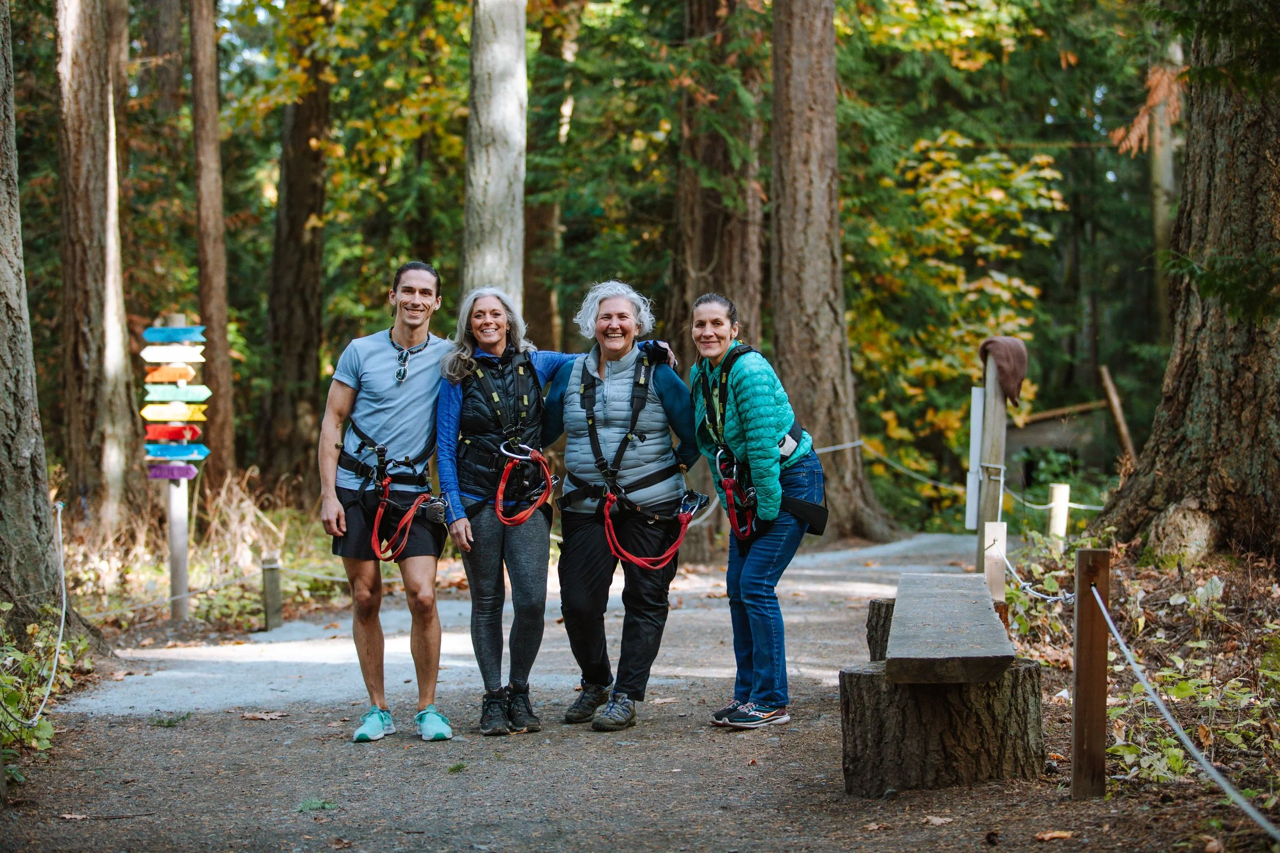 Four people, two men and two women, standing together on a wooded trail, smiling at the camera, dressed in outdoor hiking gear, with tall trees in the background.