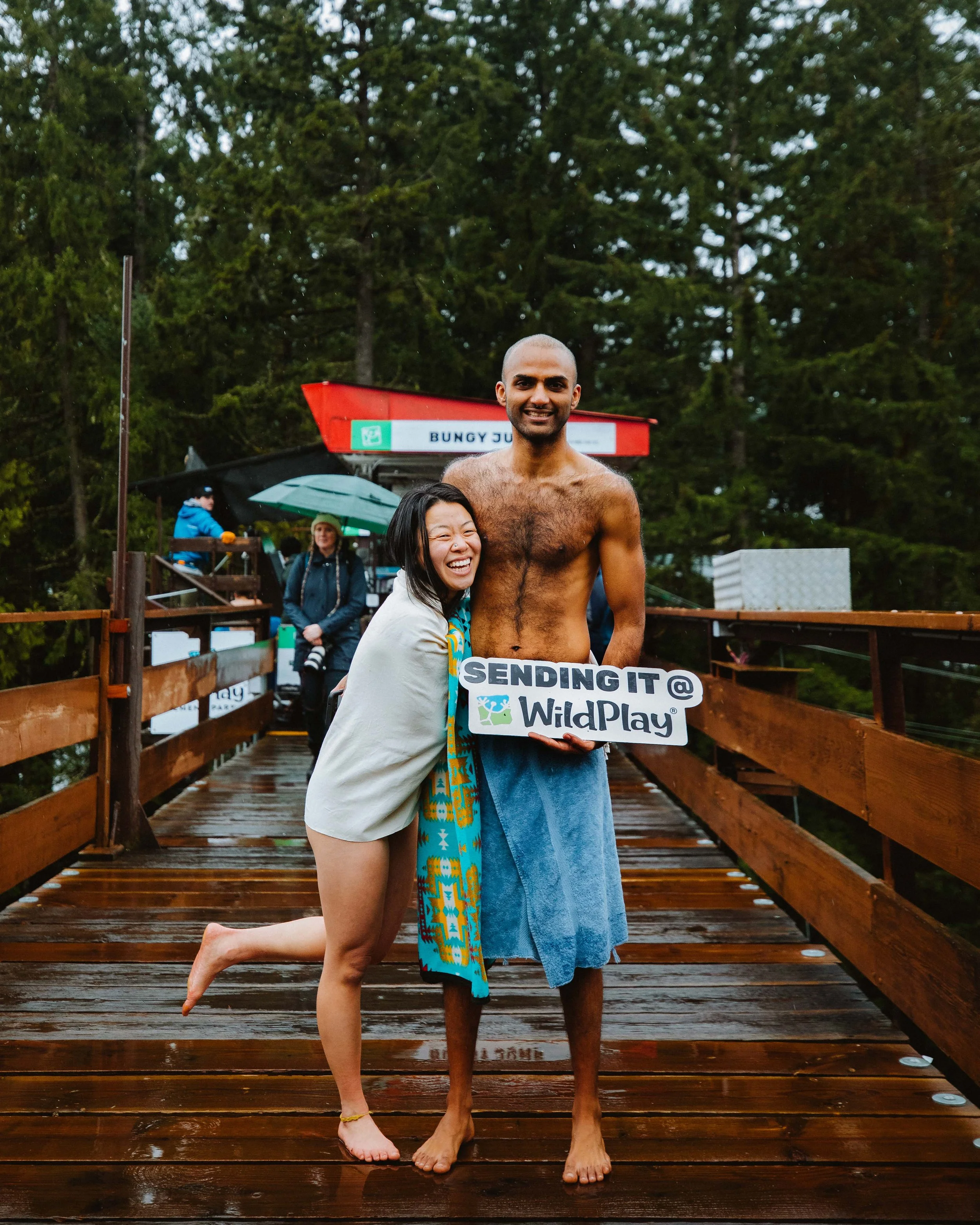 A woman and man are standing on a wooden dock, smiling and hugging. The man is shirtless and holding a sign that says 'SENDING IT @ WildPlay'. The scene appears to be outdoors with trees in the background.