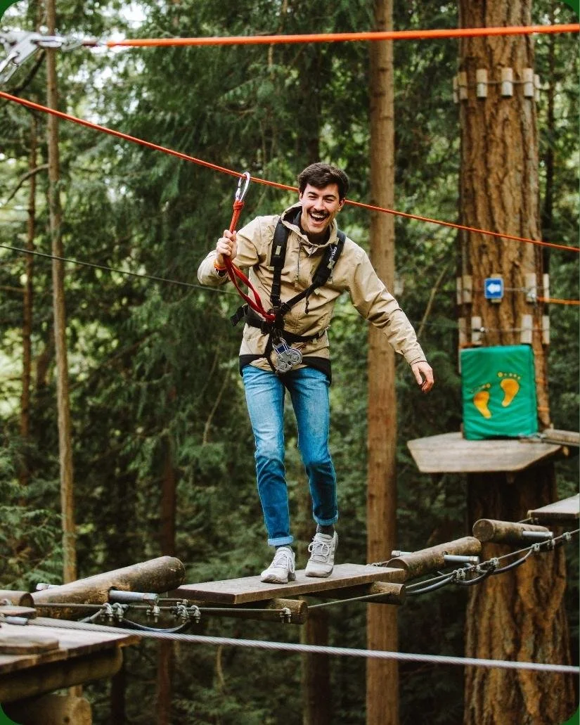 A man smiling and walking on a wooden suspension bridge at a ropes course in a forest.