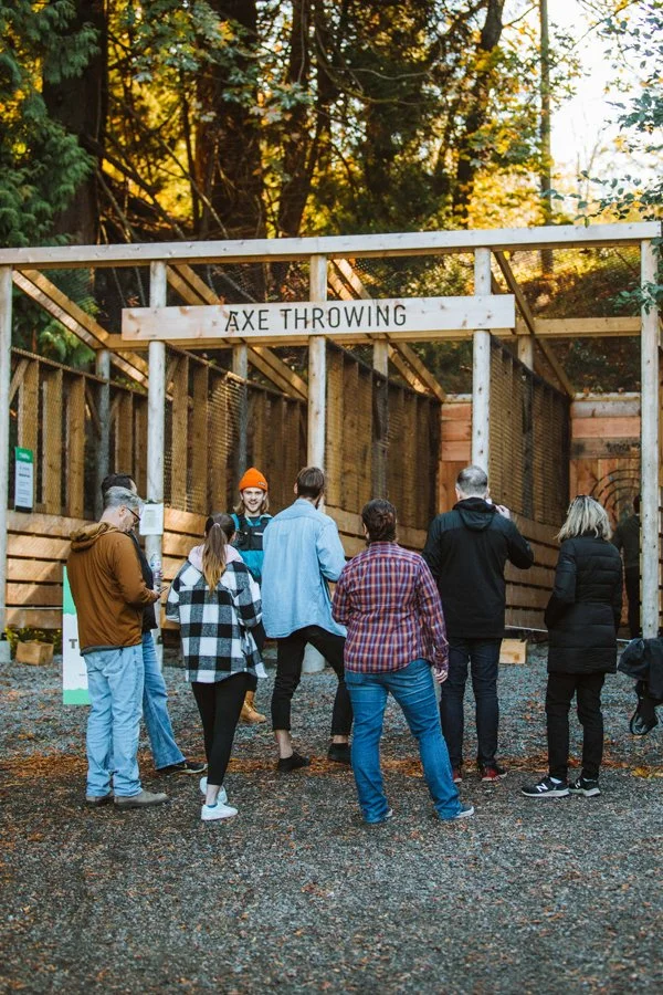 People gathered outside at an axe throwing venue, with a woman in a blue jacket and orange beanie preparing to throw an axe, under a sign that reads 'AXE THROWING', surrounded by wooden fencing and trees with autumn foliage.