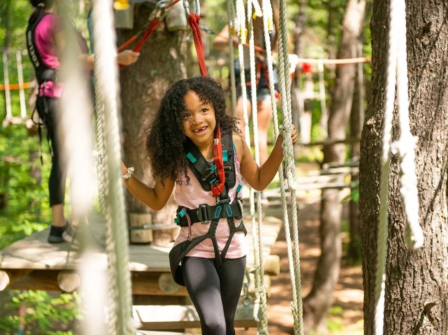 A smiling girl with curly hair navigates a rope course, wearing a harness and safety equipment, surrounded by green foliage in an outdoor adventure park WildPlay Thacher Victoria Maple Ridge Nanaimo