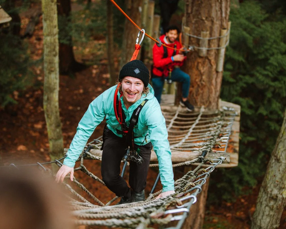 Two friends crossing a rope bridge in a forest, smiling and enjoying outdoor adventure.