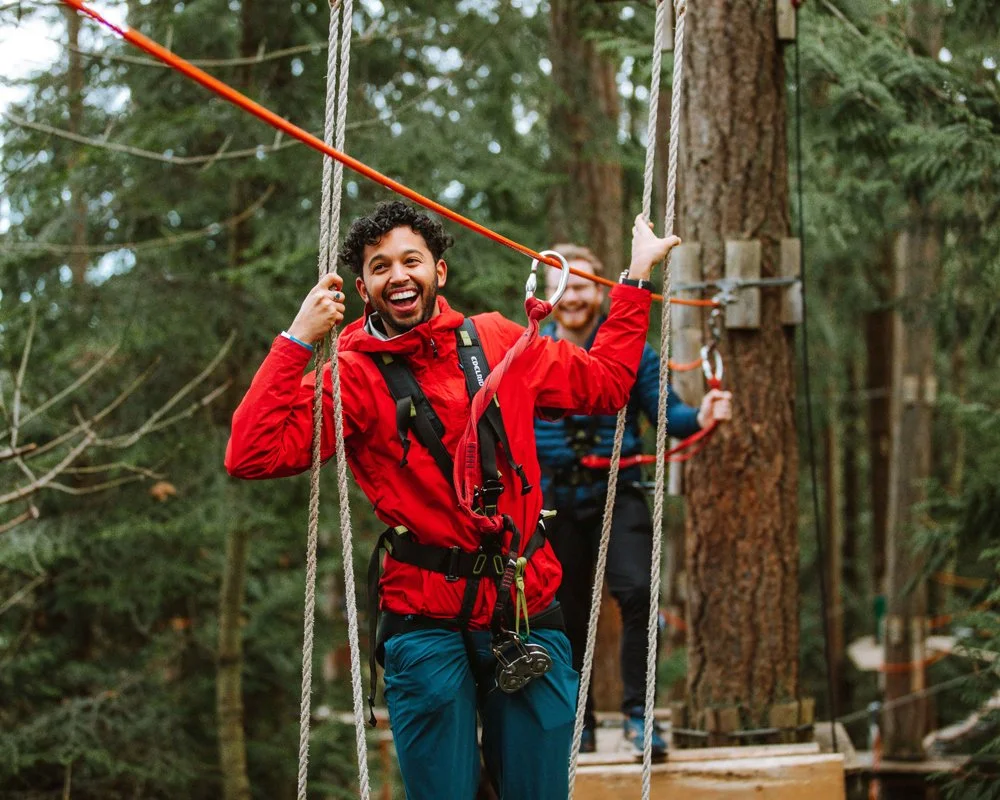 Two men on a ropes course in a forest, smiling and enjoying themselves.