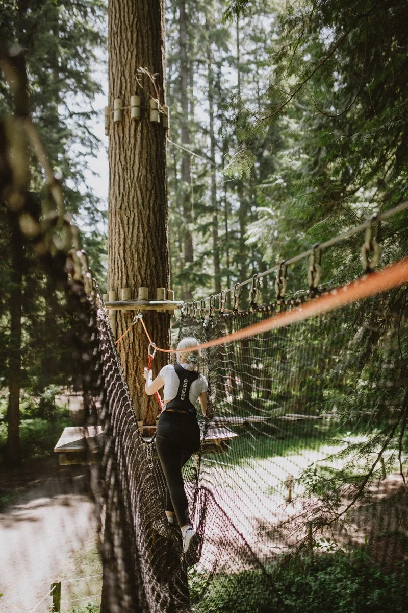 A person with a ponytail crossing a high ropes course in a forest, using safety gear and a harness.