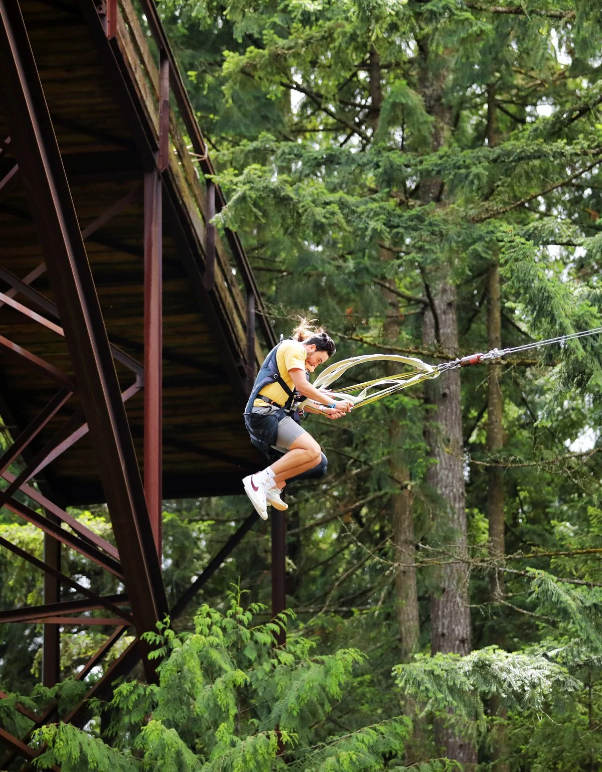 A person with long hair pulled back, wearing a yellow shirt, black shorts, and white sneakers, is zip-lining through a dense, green forest from a metal platform.