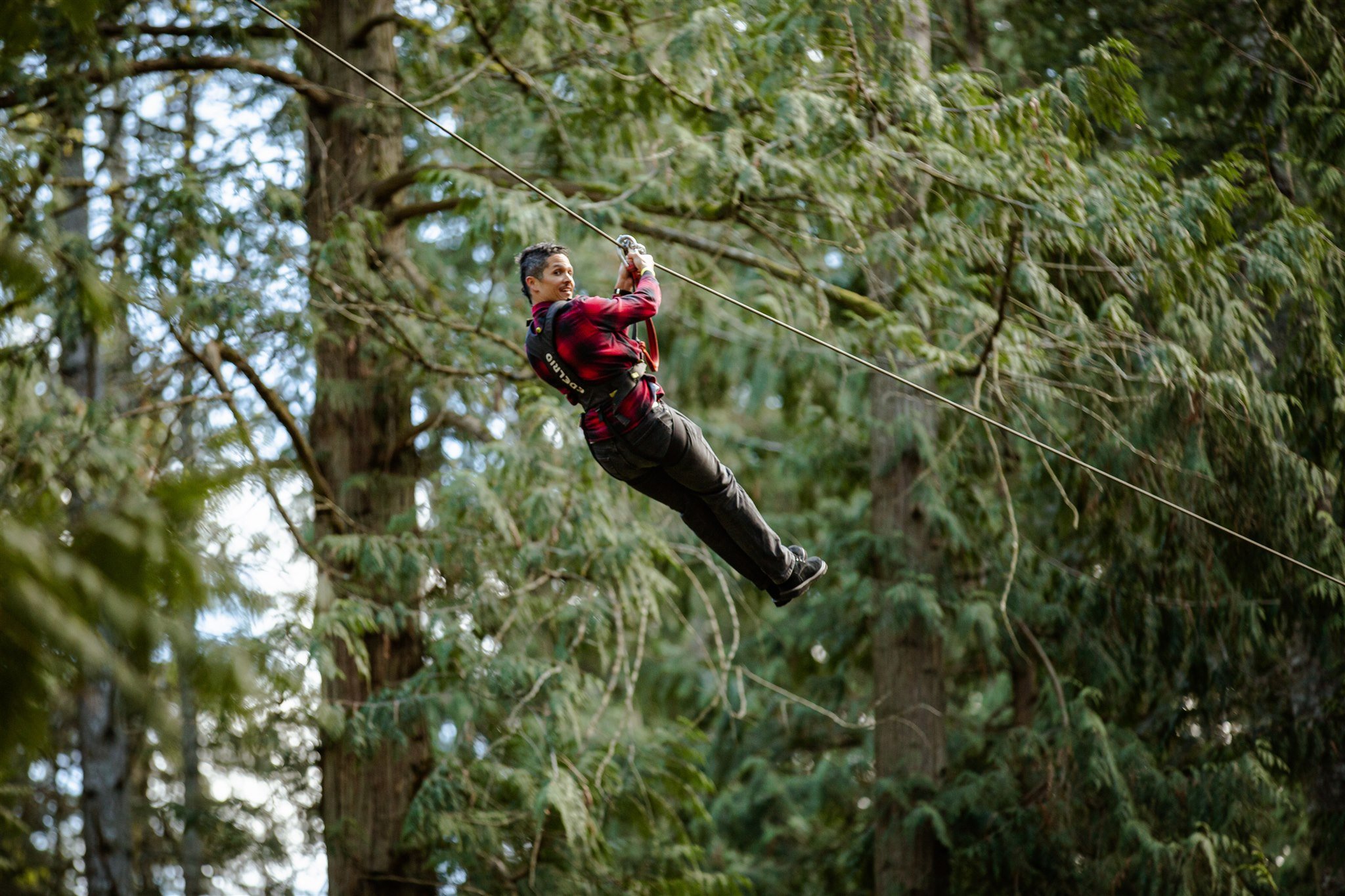 A woman wearing a red checkered shirt and black pants is ziplining through a forested area with tall trees and green foliage.