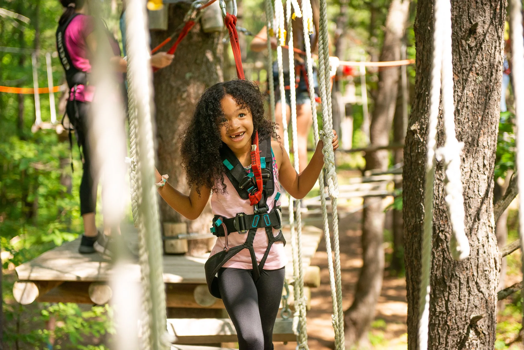 A young girl with curly hair smiling while walking on a rope bridge in a forest adventure park with other children in the background.