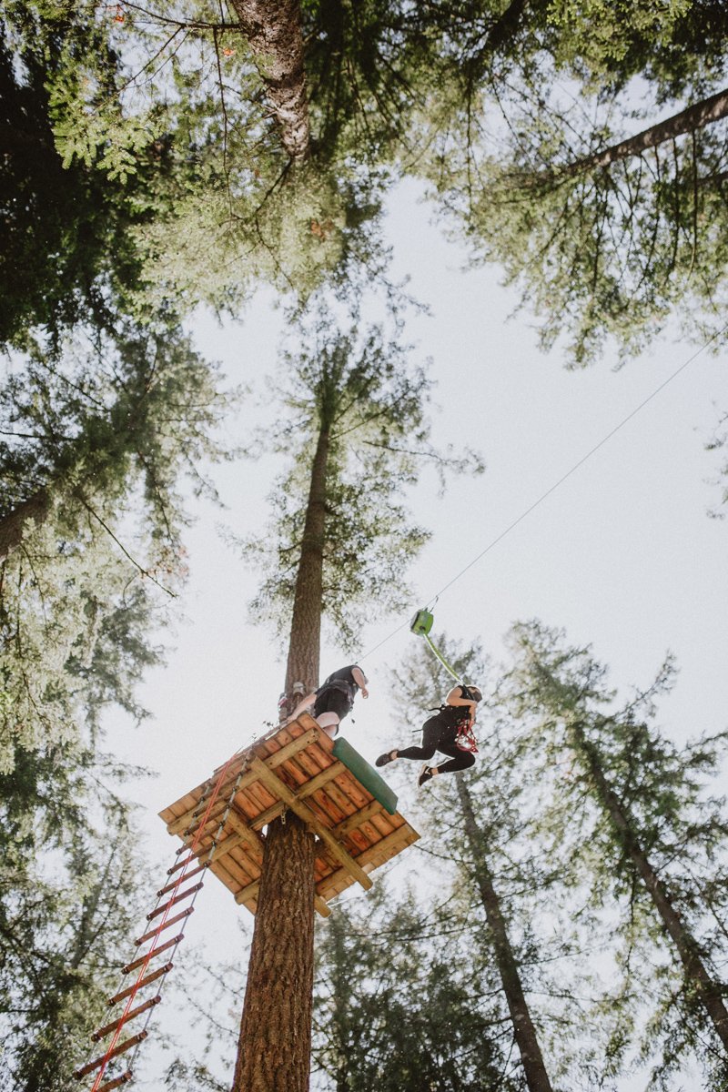 Two children on a wooden platform attached to a tall tree, playing on a zip line in a forest with tall pine trees and a partly cloudy sky.