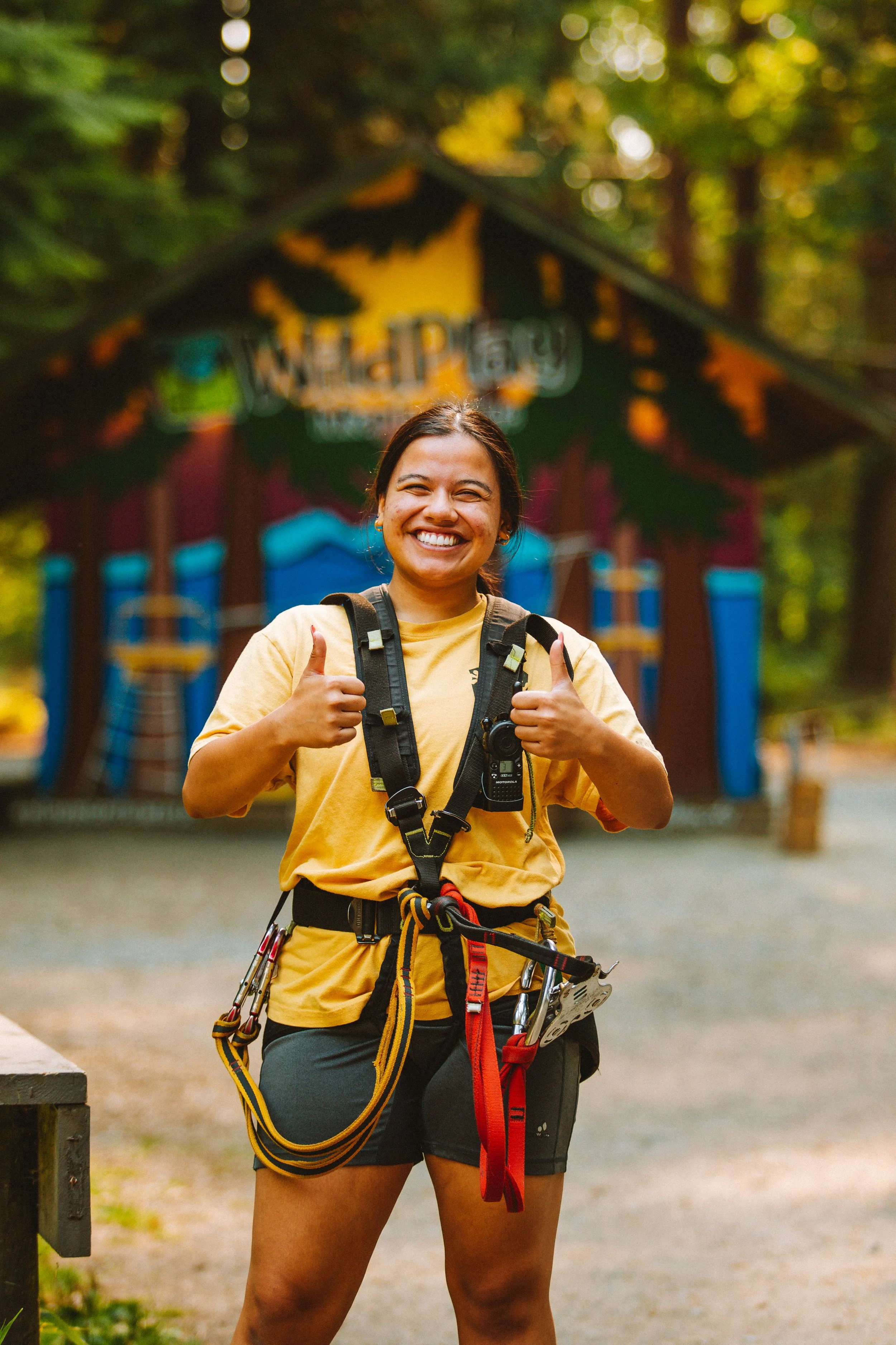 A woman in outdoor climbing gear smiling and giving two thumbs up, standing in front of a colorful wooden playground in a forested area.