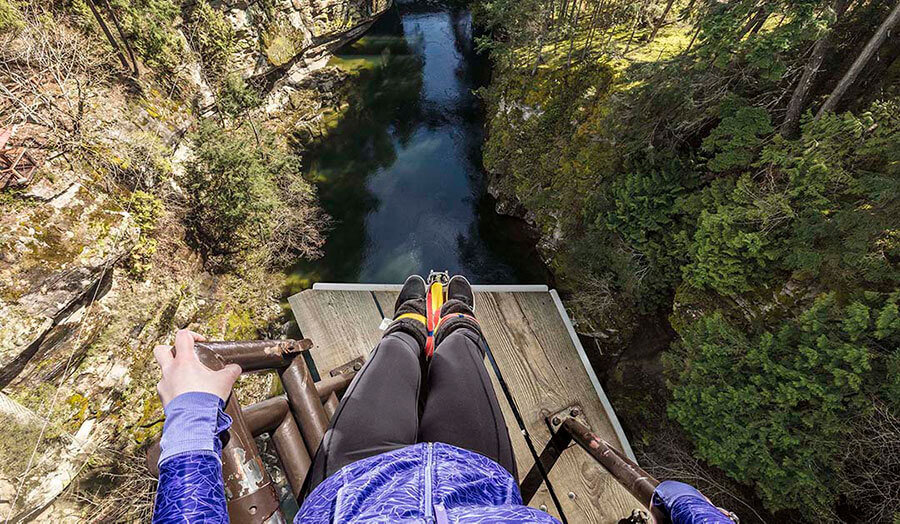 Person in blue jacket and black pants preparing to bungee jump from a platform high above a river surrounded by trees.