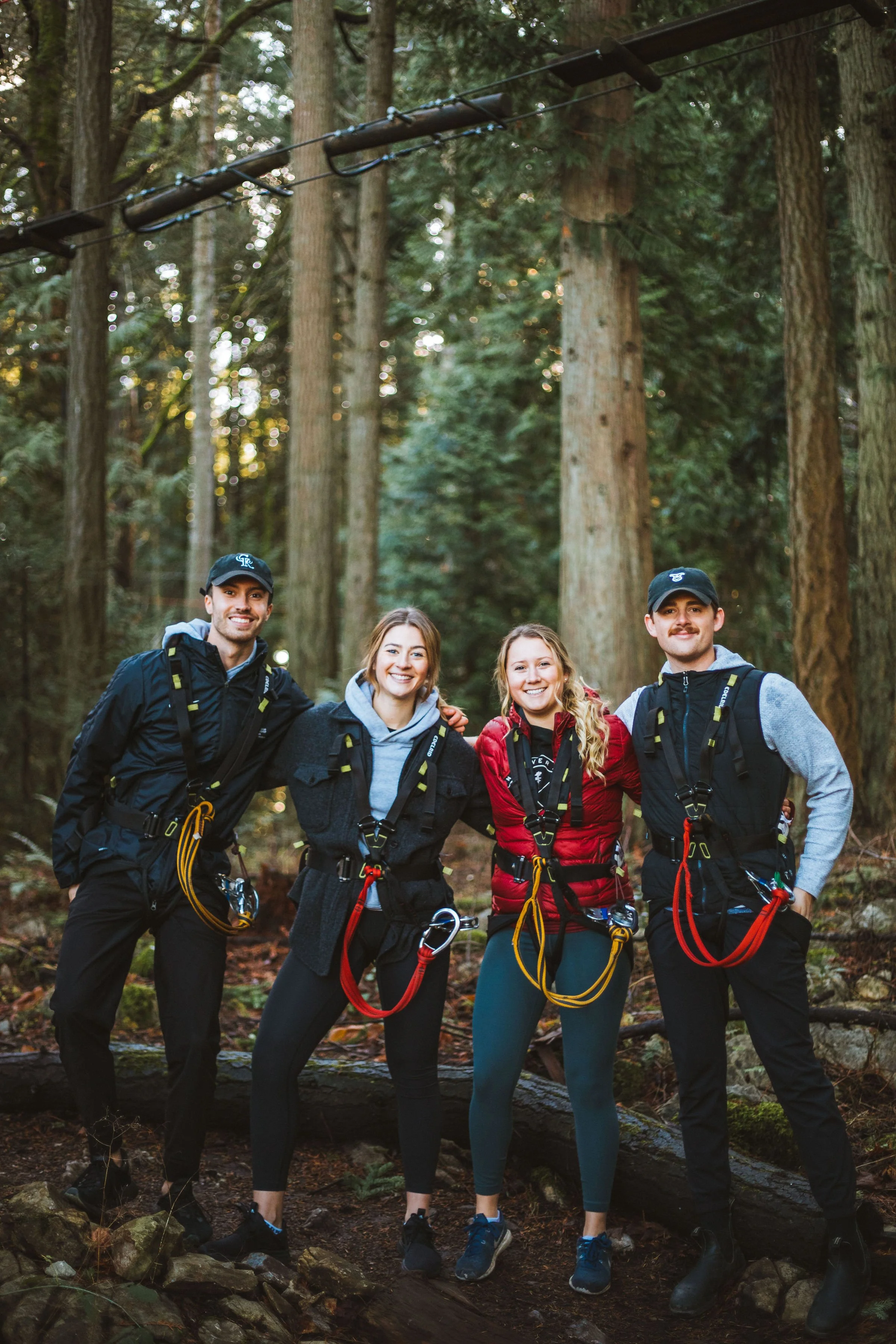 Four young friends in outdoor gear standing in a forest with tall trees, smiling and posing together after a zip-lining activity.