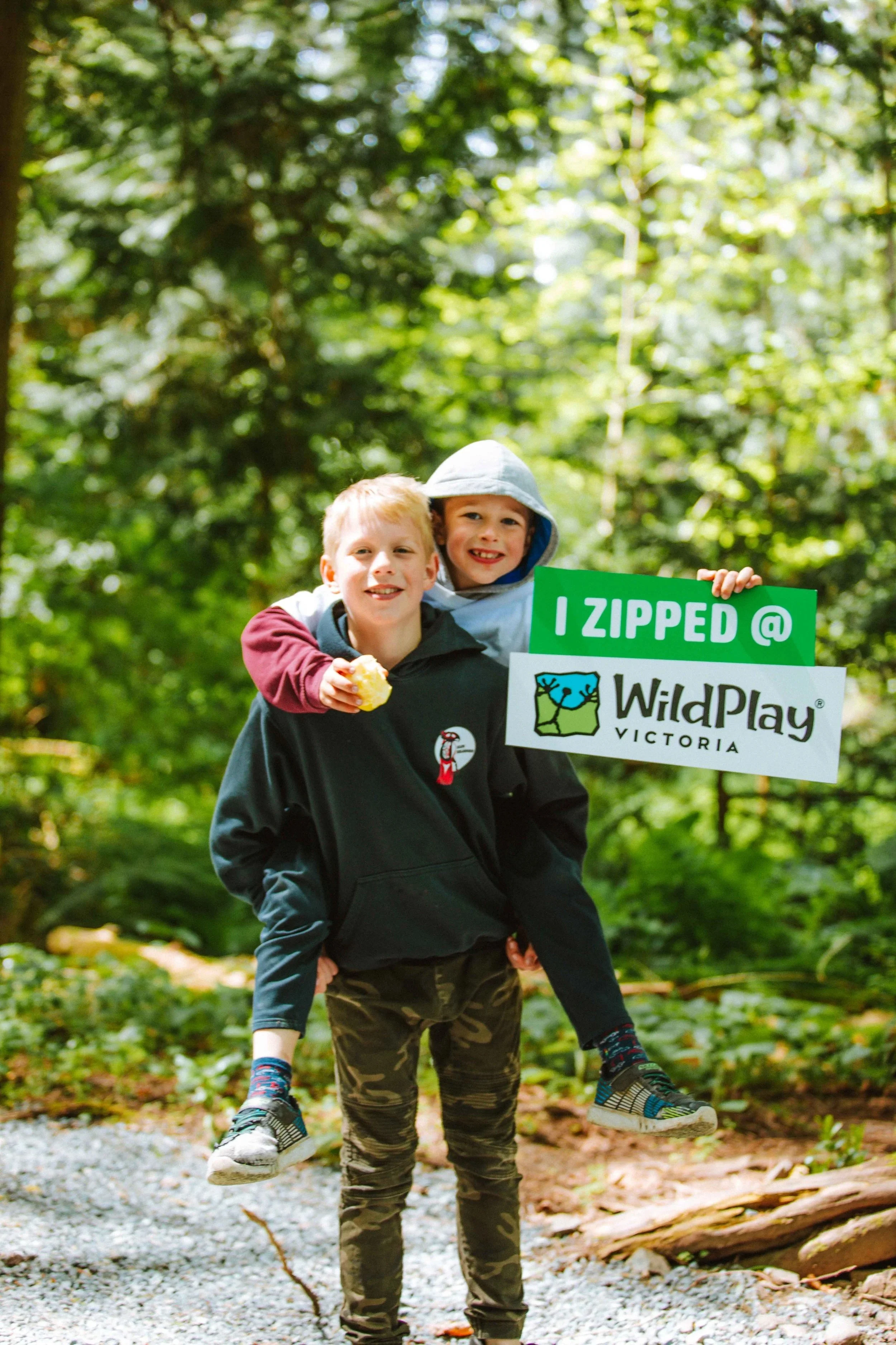 two children smiling in forest holding ‘I zipped’ sign at WildPlay Victoria adventure park after kids camp