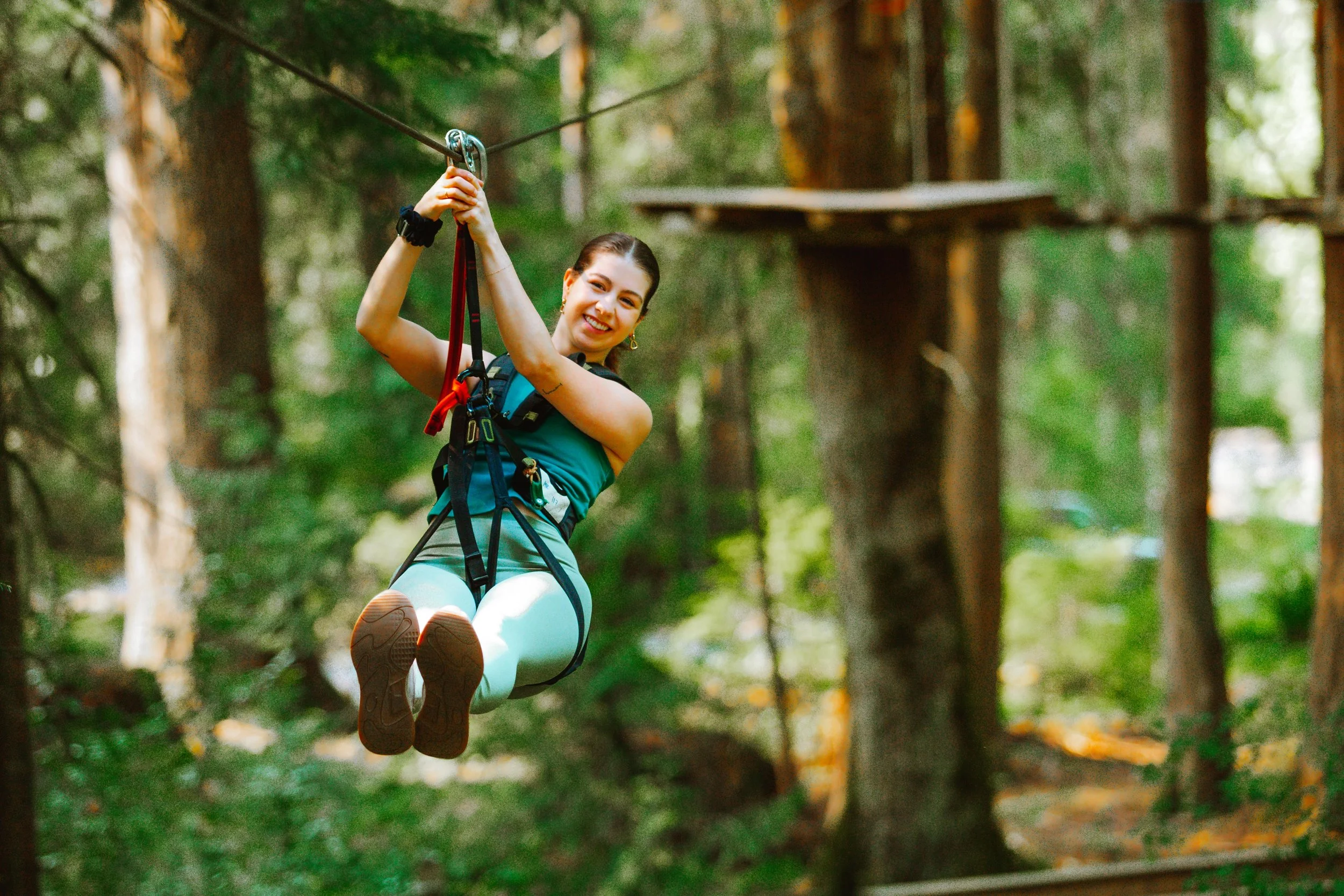 Woman enjoying a zip line adventure in a forest, smiling and holding the zip line handle while wearing safety harness and outdoor gear.