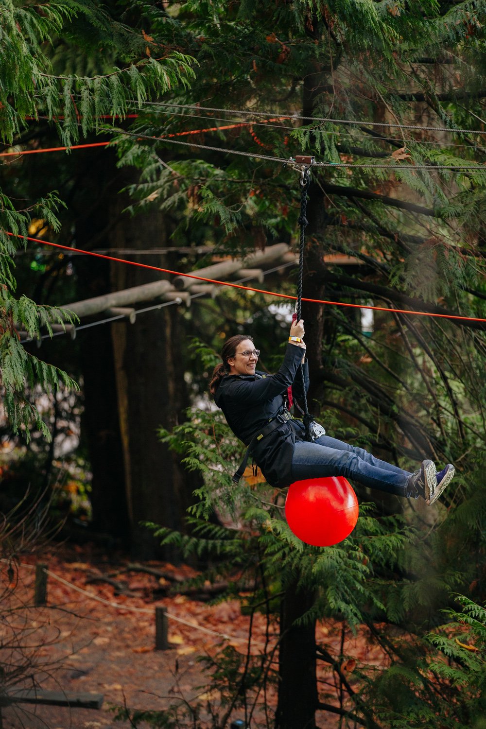 A woman riding a red ball on a zip line through a wooded area with green trees and autumn leaves on the ground.