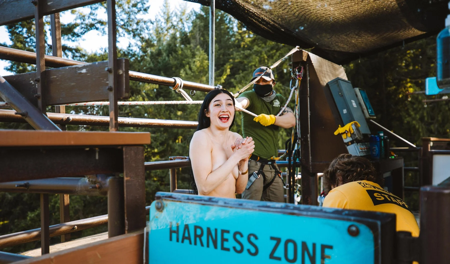 A woman with long dark hair, smiling and holding her hands together, is in a harness on a zip line. A staff member wearing a yellow shirt and a person in a green shirt and mask are assisting her. The scene takes place outdoors with trees in the backg