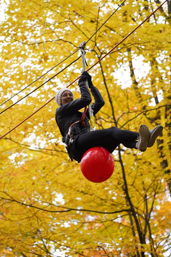 Woman riding on a large red ball attached to a zip line through autumn trees with yellow leaves.