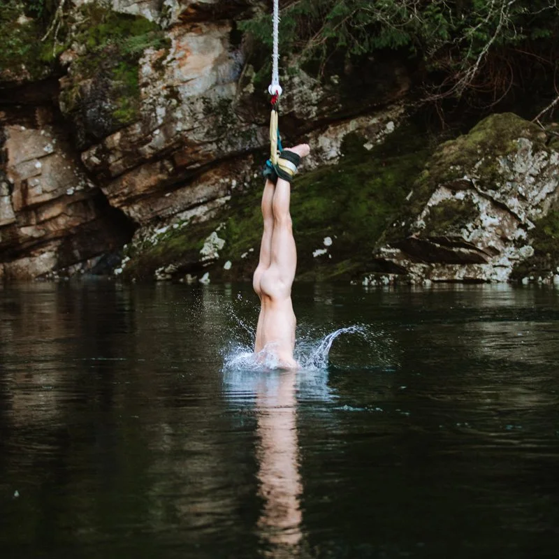 A person hanging upside down by their ankles from a rope above a body of water, with their head submerged and water splashing around.