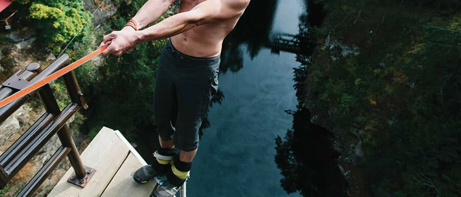 A person standing on a dock near water, preparing to jump or dive into the river below, holding a rope attached to a safety harness.