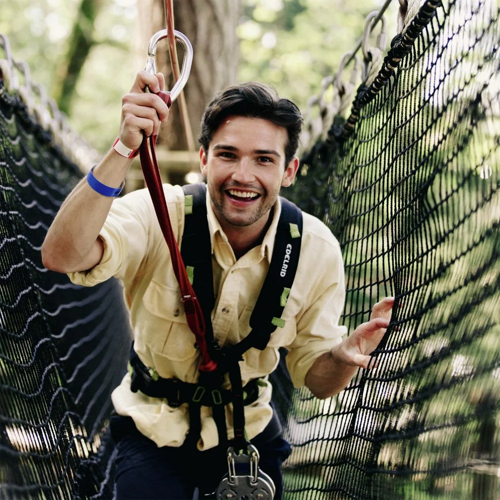 A man smiling while walking on a rope bridge in a forest, holding onto the netting with one hand and the safety harness with the other.
