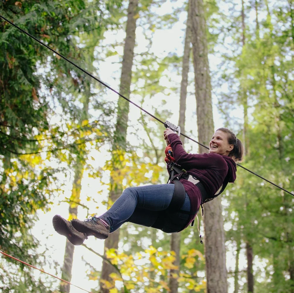 A woman smiling while ziplining through a forest with green foliage and tall trees.