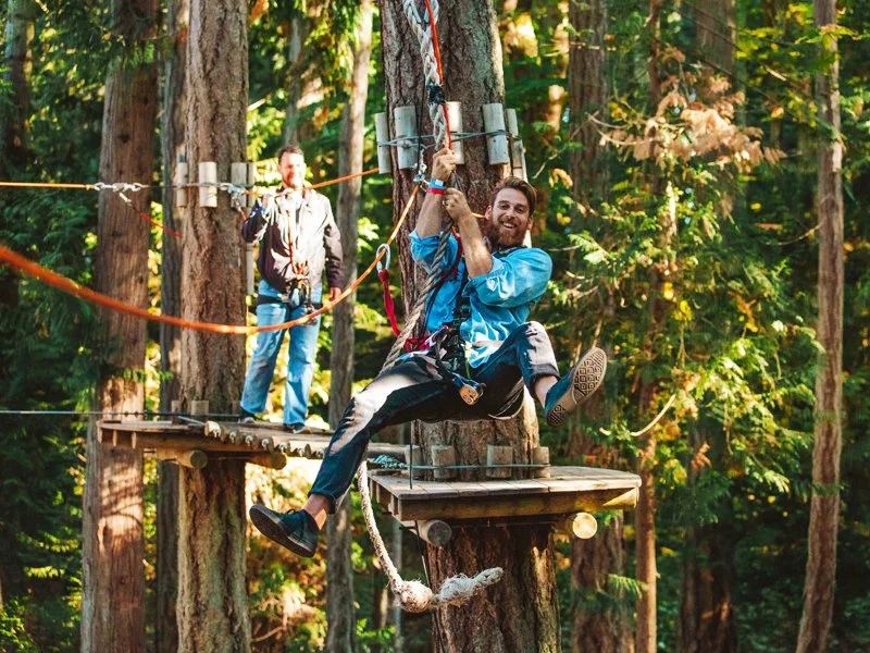 Man with a beard and blue jacket riding a zip line through a forest, smiling, with another person on a platform in the background.