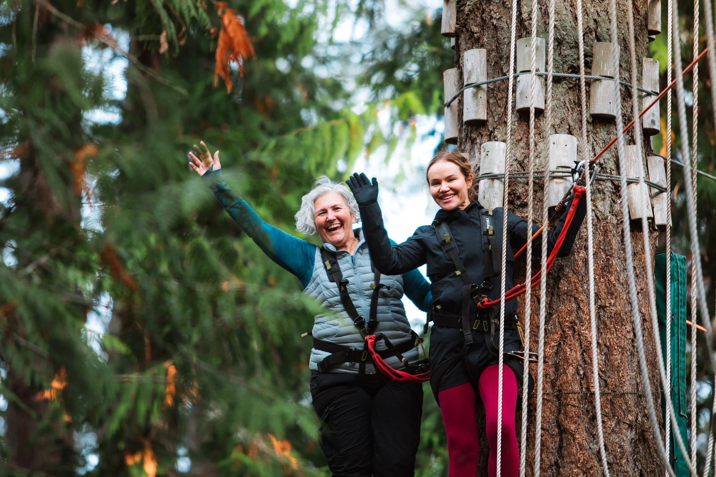 Two women smiling on a treetop adventure course, both wearing harnesses and standing on a platform at WildPlay outdoor adventure park. 