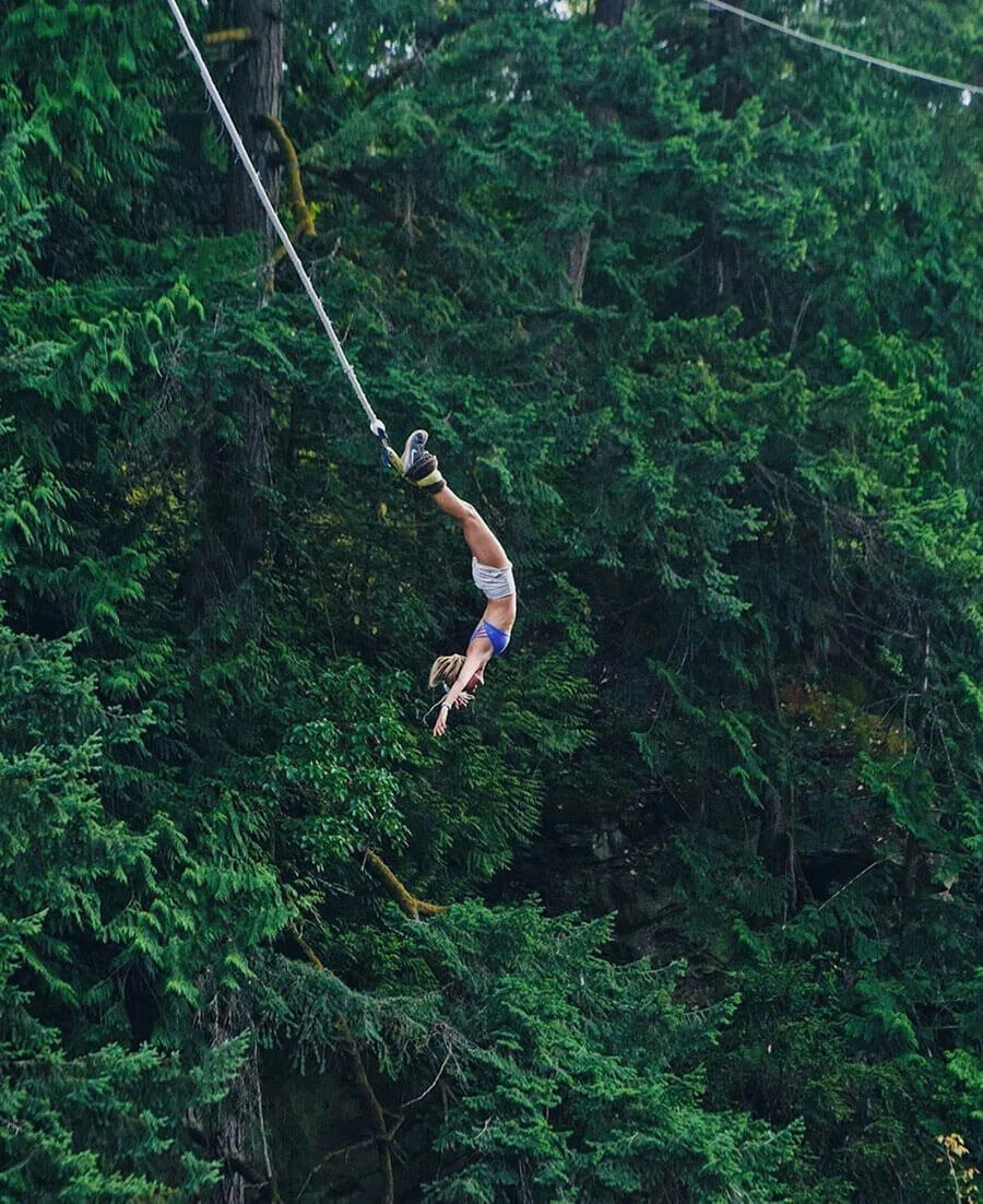 A woman is mid-air, swinging on a zip line through a green forest, with dense foliage and trees in the background.