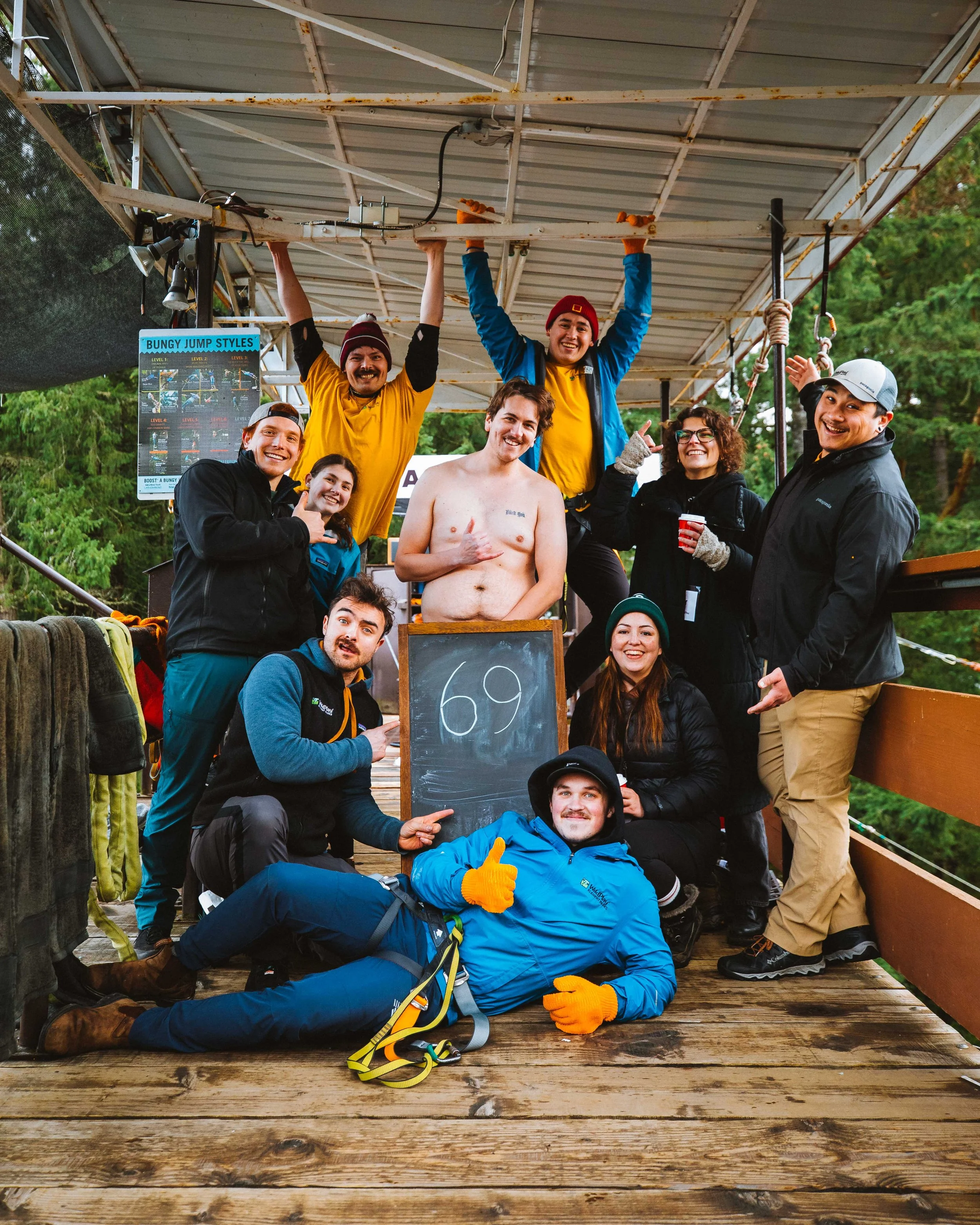 Group of friends posing on a zipline platform, some smiling and others showing thumbs up, with a chalkboard displaying the number 69, in outdoor wooded setting.