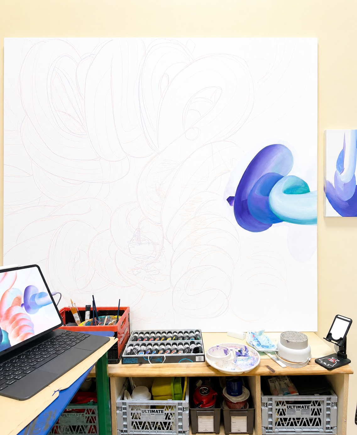 A workspace with art supplies, a laptop, and an unfinished abstract painting on the wall with blue and purple fluid shapes.