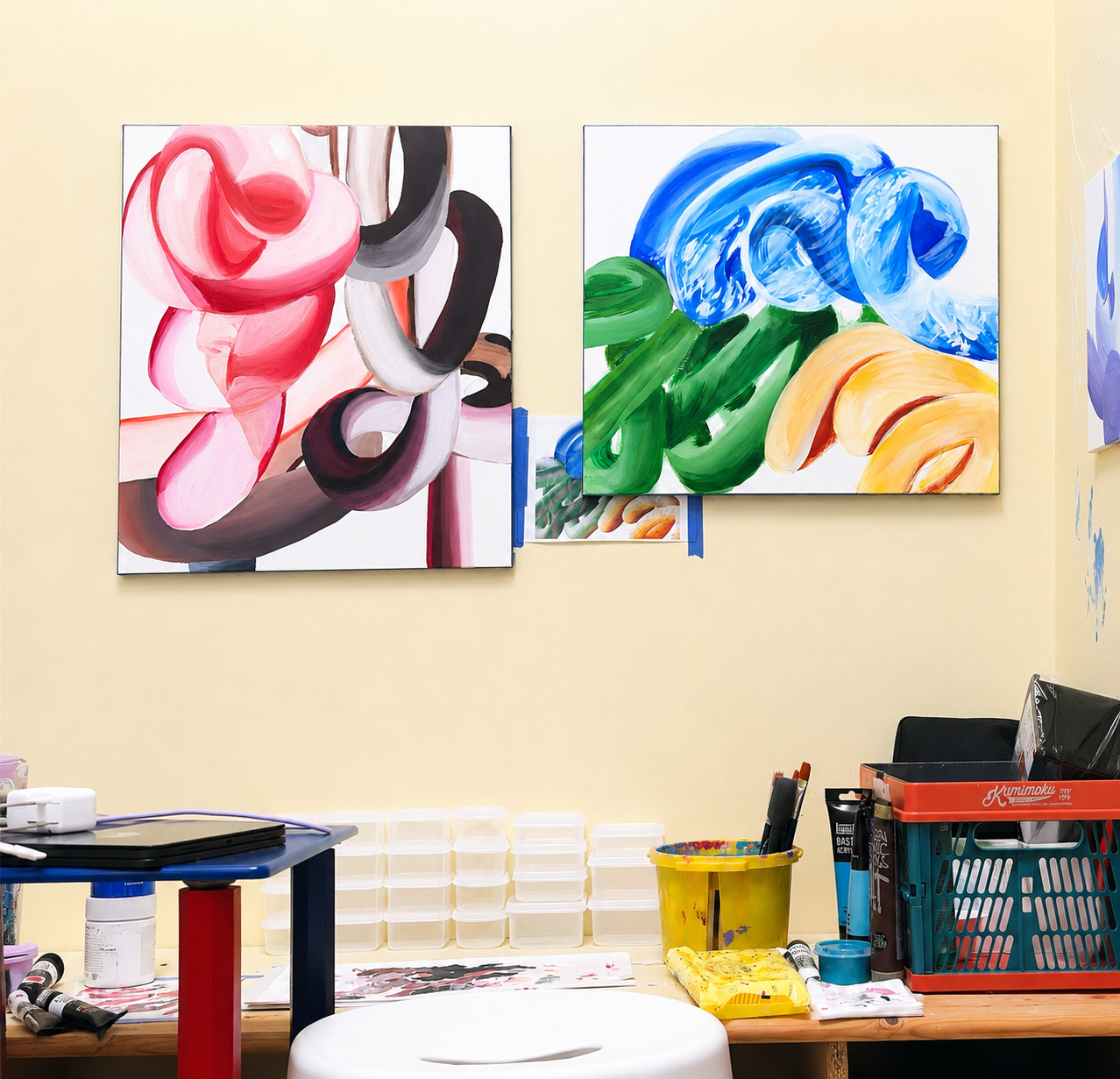 Two large abstract colorful paintings hung on a beige wall in an art studio. The left painting features pink and gray swirling shapes, while the right painting has green, blue, and yellow swirling strokes. Art supplies and storage containers are on a